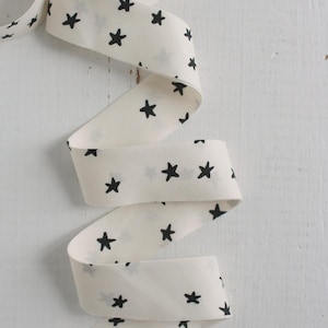 May include: Cream-colored ribbon with a repeating pattern of black stars. The ribbon is curled and draped, showcasing the star design. The background is a white, textured surface.