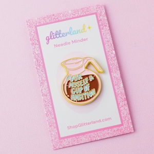 May include: A pink and gold enamel needle minder pin in the shape of a coffee pot. The pin has the text "Pour Myself a Cup of Ambition" in blue and brown. The pin is on a pink glitter card with the text "glitterland" and "Needle Minder".