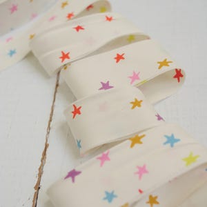 May include: Cream-colored ribbon with a repeating pattern of colorful stars in red, pink, yellow, blue, and purple. The ribbon is rolled up in a spool and unraveled, showing the star pattern. The ribbon is likely used for crafts or gift wrapping.