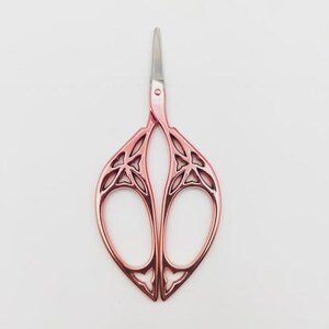May include: Rose gold embroidery scissors with a decorative floral design. The scissors have a pointed tip and a symmetrical, openwork handle. The blades are silver-colored.