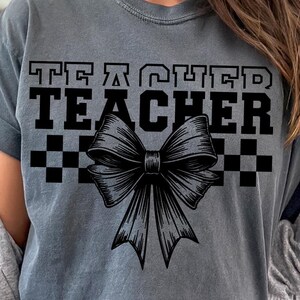 May include: A gray t-shirt with the text "Teacher" in black block letters and a black and white checkered pattern. A large black bow with a detailed design is centered below the text.