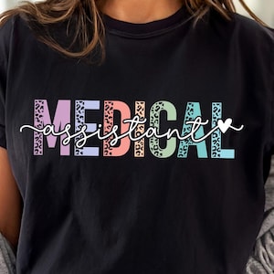 Puede incluir: Una camiseta negra con el texto "MEDICAL assistant" en colores arcoíris con un estampado de leopardo. El texto está en una fuente en negrita y juguetona con un pequeño corazón al final.