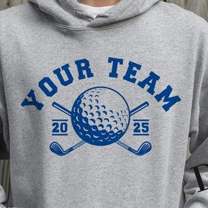 Könnte beinhalten: Ein grauer Hoodie mit einer blauen Grafik eines Golfballs und gekreuzten Golfschlägern. Der Text "YOUR TEAM" befindet sich über der Grafik, und die Zahlen "20" und "25" befinden sich auf beiden Seiten der Grafik.