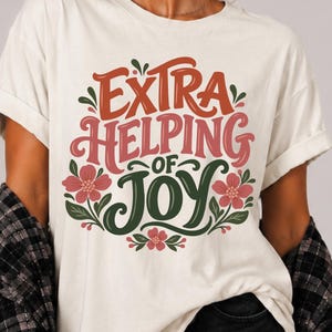 May include: An ivory t-shirt featuring the text Extra Helping of Joy in orange, pink, and dark green lettering. The design is surrounded by dark green leaves and small pink flowers. The text t-shirt colour IVORY is printed in the upper right corner.