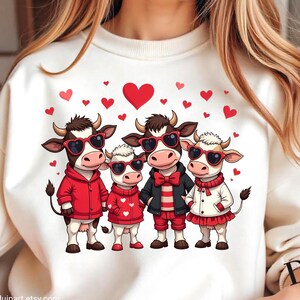 Può includere: Una felpa bianca con una grafica di quattro mucche cartoon che indossano occhiali da sole a forma di cuore rosso e vestiti rossi. Le mucche sono circondate da cuori rossi.