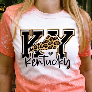 Peut inclure: Un t-shirt blanc avec un motif tie-dye orange. Le t-shirt présente un graphique noir et or des lettres "KY" avec un contour léopard de l'état du Kentucky. Le texte "Kentucky" est écrit en cursive noire sous le graphique.