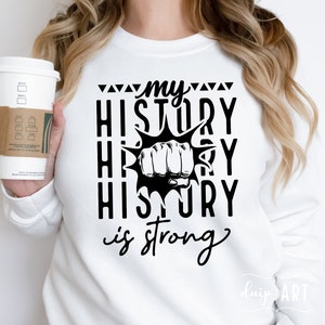 Pode incluir: Camisola branca com texto preto que diz "my history is strong" com um gráfico de punho preto no centro.
