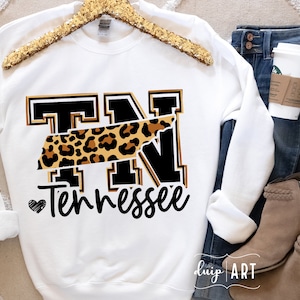 Puede incluir: Sudadera blanca con un gráfico "TN" negro y dorado con un contorno de leopardo del estado de Tennessee. El texto "Tennessee" está escrito debajo del gráfico con un pequeño corazón.