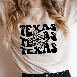 May include: A beige sweatshirt with a black graphic design of the state of Texas with a leopard print pattern and the word "Texas" repeated three times in a retro font.
