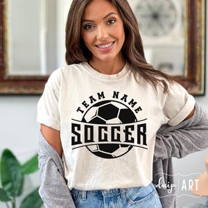 Pu&ograve; includere: Una maglietta bianca con una grafica di un pallone da calcio nero e il testo "Team Name Soccer".
