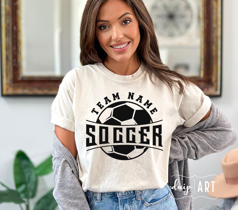 Pu&ograve; includere: Una maglietta bianca con una grafica di un pallone da calcio nero e il testo "Team Name Soccer".