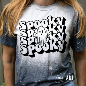 Może przedstawiać: Biała i szara koszulka z napisem "Spooky" ułożonym wielokrotnie w stylu retro. Biała grafika ducha jest wyśrodkowana w słowach. Koszulka ma wybielony, postarzany wygląd, idealny na Halloween.