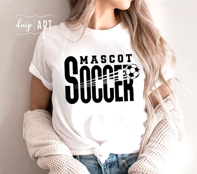 Pu&ograve; includere: Maglietta bianca con testo nero che recita "MASCOT SOCCER" e una grafica di un pallone da calcio bianco e nero.