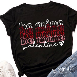 May include: Black t-shirt with the words "be mine valentine" in white, red, and pink layered text. A small white heart is next to the word "valentine". The shirt is made of a soft material.