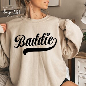 K&ouml;nnte beinhalten: Ein beiger Pullover mit dem Wort "Baddie" in schwarzer Schrift mit einem kleinen Herzen &uuml;ber dem Wort.