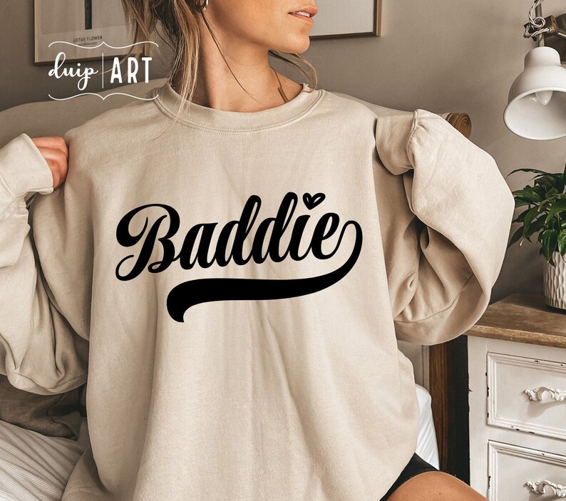 K&ouml;nnte beinhalten: Ein beiger Pullover mit dem Wort "Baddie" in schwarzer Schrift mit einem kleinen Herzen &uuml;ber dem Wort.