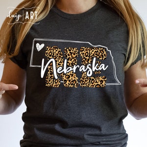 May include: A gray t-shirt with a white outline of the state of Nebraska filled with a leopard print pattern. The word "Nebraska" is written in white cursive lettering inside the state outline. A small white heart is in the top left corner of the outline.