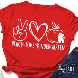 May include: Red t-shirt with a white peace sign, heart, apple, and the letter K. The text reads "Peace Love Kindergarten".