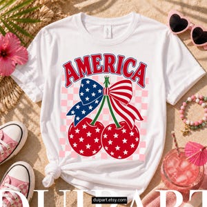 Puede incluir: Camiseta blanca con un diseño patriótico. La palabra "AMERICA" es roja, con un lazo azul con estrellas blancas y un lazo a rayas rojas y blancas. Dos cerezas con estrellas blancas están debajo. La camiseta está sobre un fondo a cuadros rosa.