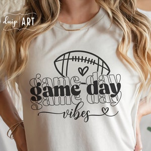 May include: White t-shirt with a black graphic design of a football and the text "game day vibes" with hearts.