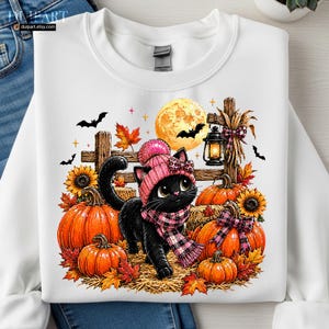 May include: A white crewneck sweatshirt featuring a graphic of a black cat wearing a pink knit hat and scarf. The cat is surrounded by orange pumpkins, sunflowers, autumn leaves, a wooden fence, a lit lantern, and a full moon with bats.