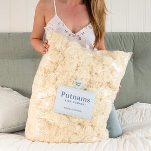 May include: A large bag of cream-coloured wool, labelled "Putnams Find Comfort Made in Devon" and "British Wool". The bag is held in front of a bed. The wool appears soft and fluffy, and is contained within a clear plastic bag.