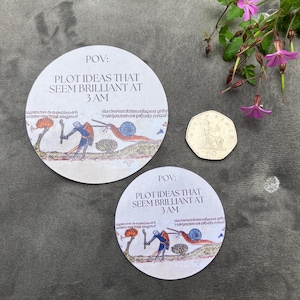 May include: Two round white stickers with a medieval illustration of a person fighting a snail. The text on the stickers reads "POV: PLOT IDEAS THAT SEEM BRILLIANT AT 3 AM".