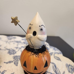 May include: A quirky Halloween ornament. A white, ghost-shaped figure sits on an orange pumpkin with a carved face. The ghost holds a gold star wand. The pumpkin has black triangle eyes and a smiling mouth. Black tinsel is around the ghost's base.