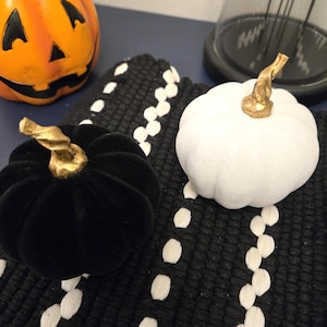 Puede incluir: Dos calabazas decorativas, una negra de terciopelo y otra blanca, con tallos dorados. Se encuentran sobre una superficie con estampado en blanco y negro. Se ve una linterna de Halloween naranja.