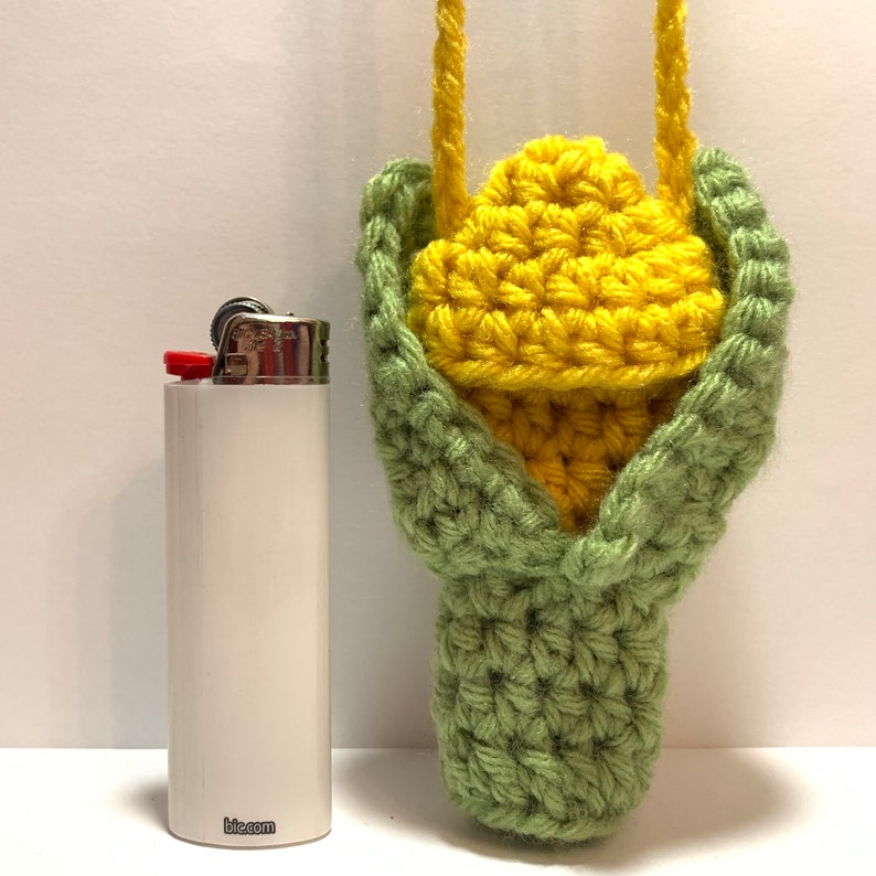 K&ouml;nnte beinhalten: Ein geh&auml;kelter Maiskolben mit gr&uuml;nen H&uuml;llen und einem gelben Kolben. Der Mais h&auml;ngt an einer Schnur und befindet sich neben einem wei&szlig;en Feuerzeug mit dem Aufdruck "bic.com".
