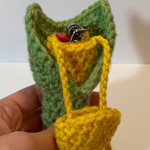 K&ouml;nnte beinhalten: Eine geh&auml;kelte gr&uuml;ne und gelbe Tasche mit einem kleinen Feuerzeug im Inneren. Die Tasche hat einen Riemen und ist so konzipiert, dass sie einen kleinen Gegenstand aufnehmen kann.