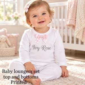 May include: A white long-sleeved top and matching trousers set featuring a pink satin bow at the neckline and the name Ivy Rose printed in grey script. The outfit includes a coordinating pink hair bow accessory for a complete look.