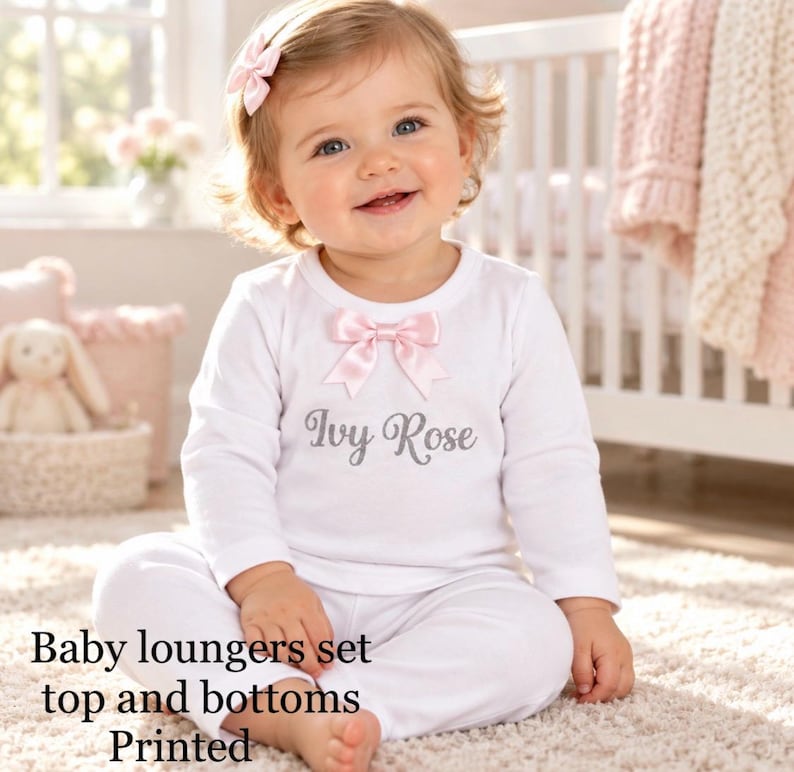 May include: A white long-sleeved top and matching trousers set featuring a pink satin bow at the neckline and the name Ivy Rose printed in grey script. The outfit includes a coordinating pink hair bow accessory for a complete look.