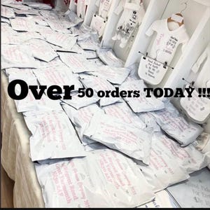 May include: A large collection of white shipping mailers stacked on a table, each printed with pink text including the address 14 Regent Road, Great Yarmouth, NR30 2AF. White baby clothing items with printed text hang on a rack in the background.