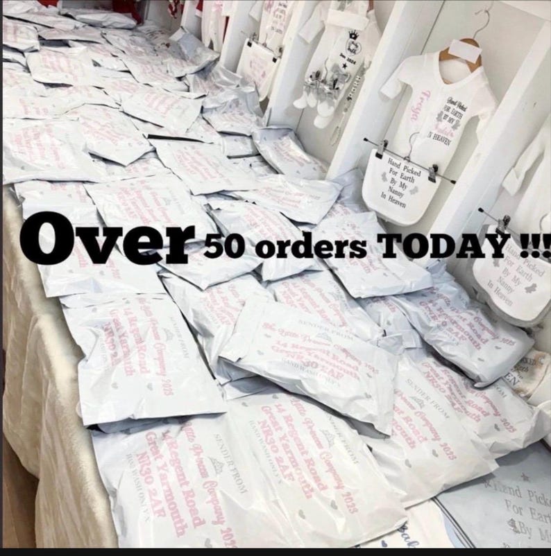 May include: A large collection of white shipping mailers stacked on a table, each printed with pink text including the address 14 Regent Road, Great Yarmouth, NR30 2AF. White baby clothing items with printed text hang on a rack in the background.