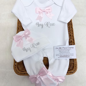 May include: A white long-sleeved baby bodysuit and matching hat set featuring the name Ivy Rose embroidered in grey. The outfit is decorated with light pink satin bows on the chest, the hat, and the feet of the bodysuit.