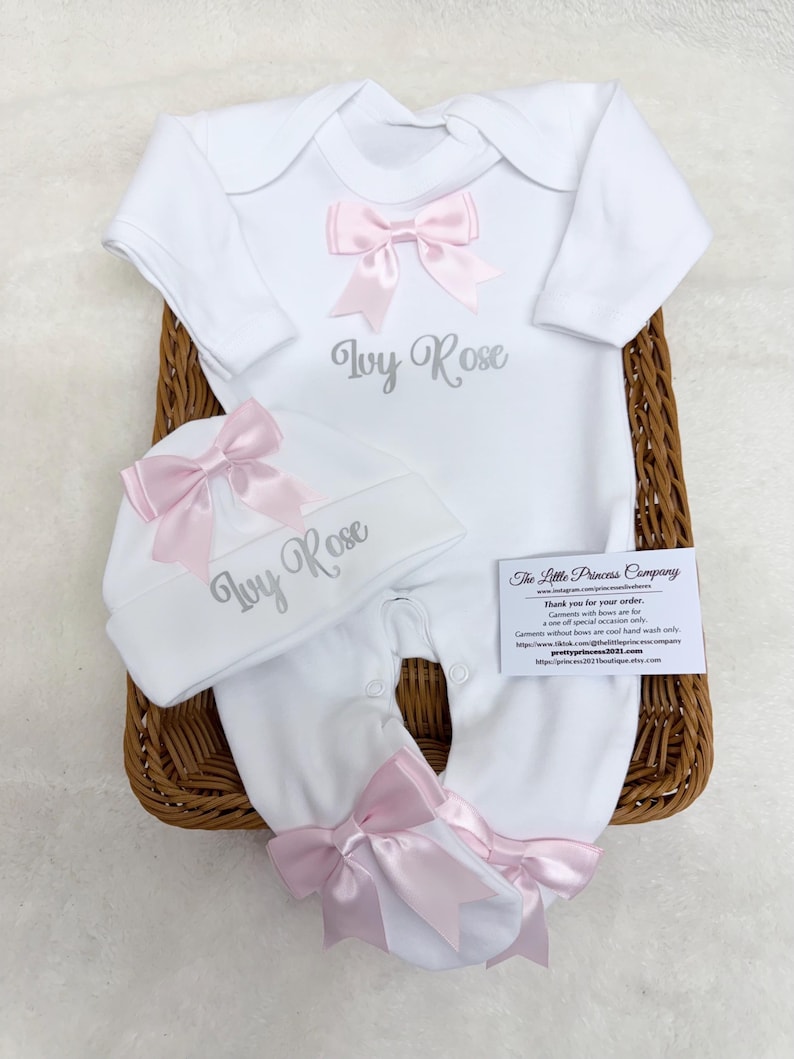 May include: A white long-sleeved baby bodysuit and matching hat set featuring the name Ivy Rose embroidered in grey. The outfit is decorated with light pink satin bows on the chest, the hat, and the feet of the bodysuit.