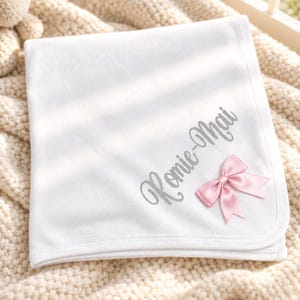 May include: A white baby blanket featuring the embroidered name Romie-Mai in a silver script font. The blanket is adorned with a small, light pink satin ribbon bow attached to the lower right corner, adding a subtle decorative accent.