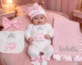 May include: A white long-sleeved baby bodysuit featuring a silver crown and the text Princess Isabella Has Arrived, a matching white hat with a pink bow and the name Isabella, a white bib with a crown and bow, and a pink textured blanket with the name Isabella.
