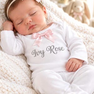 May include: A white long-sleeved baby bodysuit featuring a light pink satin bow at the neckline and the name Ivy Rose printed in a grey script font across the chest. The garment is worn by a sleeping infant with a matching pink headband.