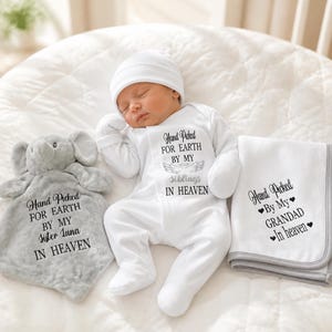 May include: A white long-sleeved baby onesie with a matching beanie, a grey elephant-shaped security blanket, and a white blanket with grey trim. The onesie reads Hand Picked FOR EARTH BY MY siblings IN HEAVEN, the blanket reads Hand Picked FOR EARTH BY MY SISTER LUNA IN HEAVEN, and the white blanket reads Hand Picked By My GRANDAD In heaven.