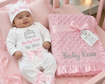 May include: A white long-sleeve baby bodysuit with a pink bow, a matching white beanie with the text Baby Kaur, and a pink dimpled baby blanket with the name Baby Kaur embroidered in silver. The bodysuit features a crown and the text Princess Baby Kaur Has Arrived.