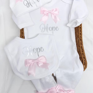 May include: A white baby outfit set including a long-sleeve footed sleeper, a bib, and a hat. Each piece features the name Hope in silver script and is decorated with pink satin bows. A small white card with black text is included.