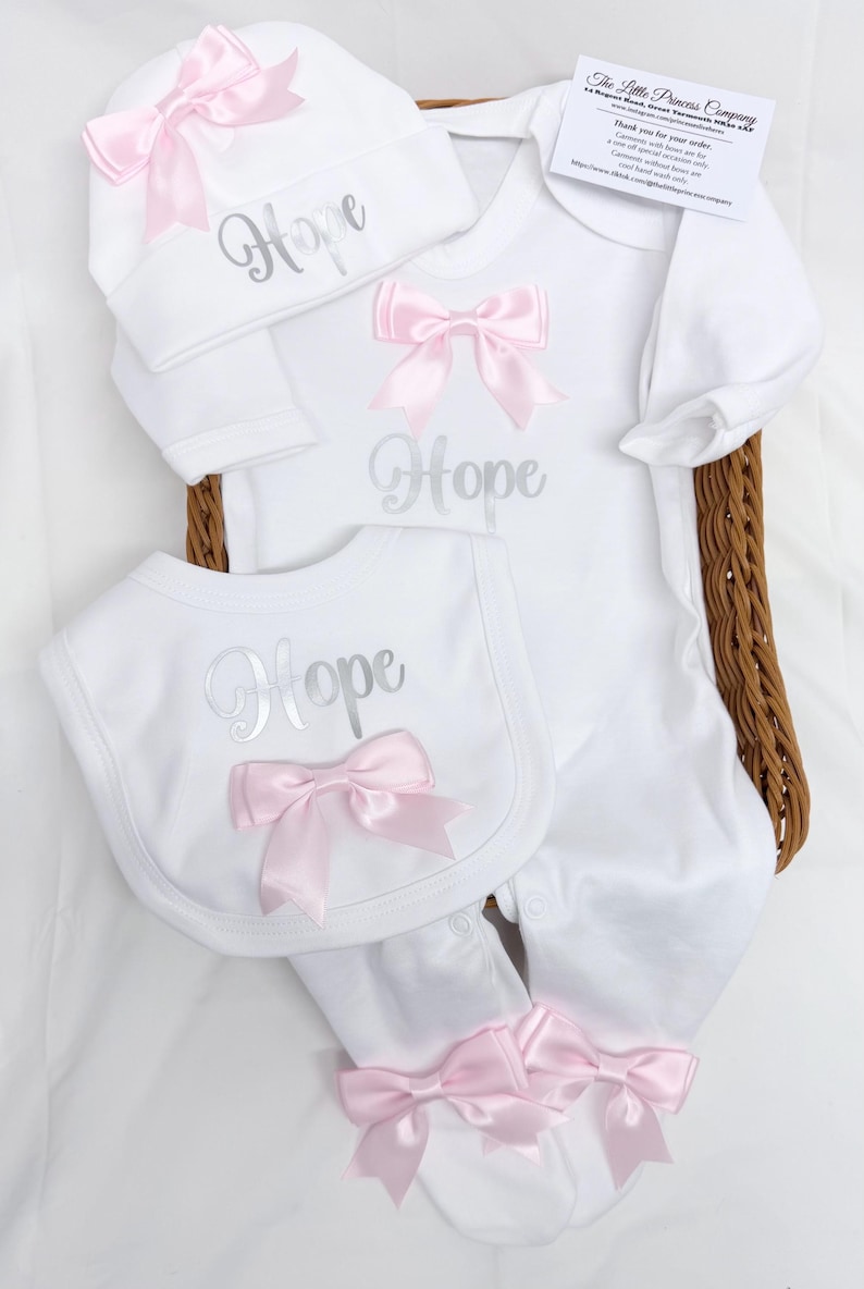 May include: A white baby outfit set including a long-sleeve footed sleeper, a bib, and a hat. Each piece features the name Hope in silver script and is decorated with pink satin bows. A small white card with black text is included.