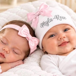 May include: A white baby hat with the embroidered name Myla-Rose in silver thread and a light pink satin bow, paired with a matching light pink satin bow headband. Both items are made of soft fabric and feature a coordinated design.