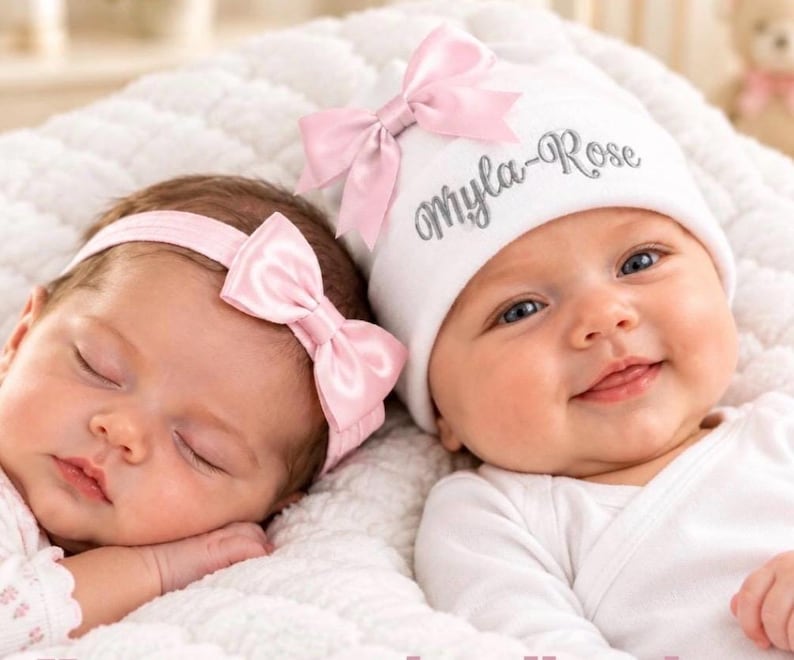 May include: A white baby hat with the embroidered name Myla-Rose in silver thread and a light pink satin bow, paired with a matching light pink satin bow headband. Both items are made of soft fabric and feature a coordinated design.