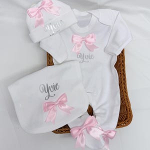 May include: A white baby clothing set including a long-sleeve footed sleepsuit, a beanie hat, and a bib. Each piece features a pink satin bow and the name Yvie embroidered in silver script. The items are arranged together in a brown woven basket.