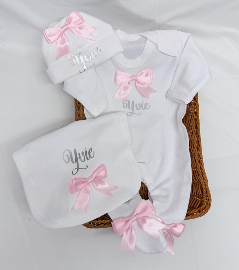 May include: A white baby clothing set including a long-sleeve footed sleepsuit, a beanie hat, and a bib. Each piece features a pink satin bow and the name Yvie embroidered in silver script. The items are arranged together in a brown woven basket.