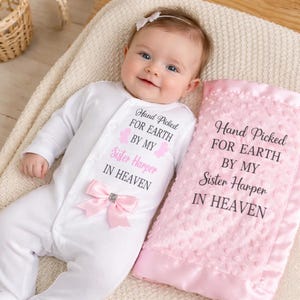 May include: A baby in a white onesie with a pink bow and the text "Hand Picked For Earth By My Sister Harper In Heaven." A pink blanket with the same text is next to the baby.