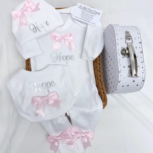 May include: A white baby set including a long-sleeve footed sleepsuit, a bib, and a hat, all featuring the name Hope in silver script and light pink satin bows. The set is presented in a wicker basket with a white suitcase featuring silver stars.